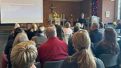 Eine Leinwand, ein Vortragender und in mehreren Sitzreihen sitzen zahlreiche Menschen. Sie verbindet, dass sie alle in Verantwortung für evangelische Kitas im Kirchenkreis Rantzau-Münsterdorf stehen.