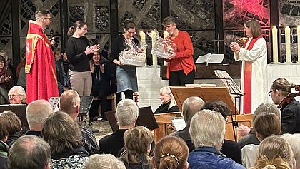 Pastorin Britta Stender und Pastorin Corinna Schmucker stehen in der Lutherkirche in Elmshorn und nehmen Geschenke entgegen, umgeben von Gemeindemitgliedern. Im Hintergrund ist ein großes, beleuchtetes Kirchenfenster zu sehen. Die Kirche ist gut gefüllt. Pastor Christofer Klaas steht links im Bild in liturgischem Gewand.