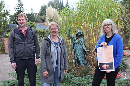 Drei Personen zeigen vor einer Engel-Statue auf dem Friedhof ein Plakat
