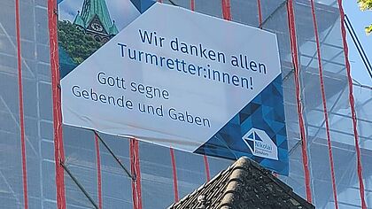 Ein Banner mit Dankesworten hängt an einem Gerüst des Kirchturms.