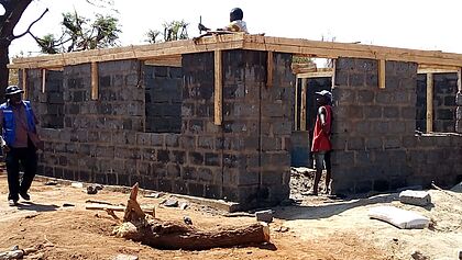 Rohbau eines Hauses in Kenia. Das Erdgeschoss ist errichtet, ein Arbeiter baut das 1. mit Holzscheiten an. Zwei weitere Männer im Bild. 