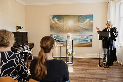 Zu sehen ist der Raum der Stille. Das Wandbild zeigt Wasser und licht. Alles ist in blau und brauntönen gehalten. Pastorin Warnke feiert in dem Raum eine Andacht. Der Raum steht allen Konfessionen offen.