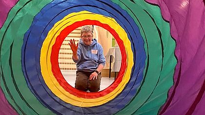Blick durch einen bunten Spieltunnel mit konzentrischen farbigen Ringen. Am anderen Ende des Tunnels sitzt eine Person in einem hellblauen Hoodie auf dem Boden und hebt eine Hand, als würde sie winken oder ein Zeichen geben. Hinter der Person sind helle Raumstrukturen erkennbar.