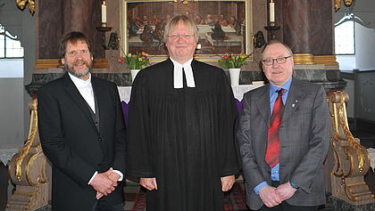 Auf dem Foto ist Pastor Lempelius zusammen mit den Kirchengemeinderatsvorsitzenden aus St. Margarethen, Achim Franzen, und Brokdorf, Michael Dombrowski, zu sehen. Sie stehen vor dem Altar in der Kirche zu St. Margarethen.