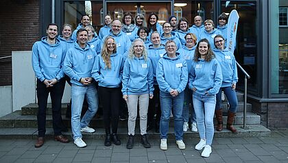 Das Team Kita-Werk steht vor dem Kirchlichen Zentrum Elmshorn. Alle tragen blaue Hoodies mit dem Logo des Kirchenkreises Rantzau-Münsterdorf.