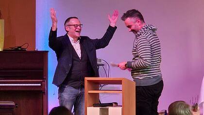 Rolf Paulsen war Religionslehrer am RBZ in Itzehoe. Hier zu sehen ist er mit Propst Steffen Paar. Er reist die Arme hoch vor Erleichterung im Moment seiner Entpflichtung durch den Propst.