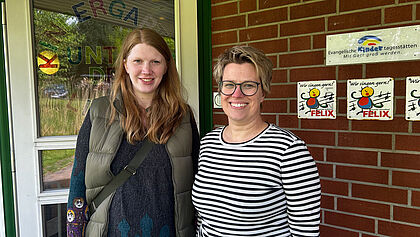 Zwei Frauen stehen vor dem Eingang der Evangelischen Kita Unter den Linden in Oelixdorf. Im Hintergrund ist eine Tür und eine Backsteinwand zu sehen. Auf der Wand hängen Siegel, die das evangelische Profil und einen Schwerpunkt auf Gesang bescheinigen.