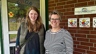 Zwei Frauen stehen vor dem Eingang der Evangelischen Kita Unter den Linden in Oelixdorf. Im Hintergrund ist eine Tür und eine Backsteinwand zu sehen. Auf der Wand hängen Siegel, die das evangelische Profil und einen Schwerpunkt auf Gesang bescheinigen.