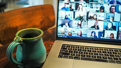 Aufgeklappter Laptop mit Videokonferenzkacheln auf dem Bildschirm und einer Tasse