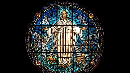 Zu sehen ist ein detailreiches Kirchenfenster, auf dem vorne Jesus Christus zu sehen ist, der seine Arme ausbreitet.