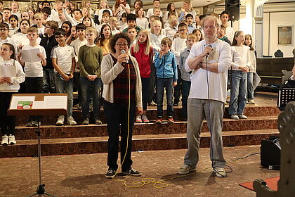 Die beiden Kantor:innen Landmesser und Reinke stehen in der St. Laurentii-Kirche. Hinter ihnen der Kinderchor.