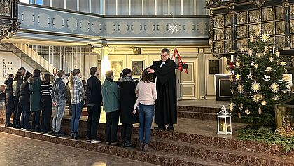 Propst Thielko Stadtand segnet eine Mitarbeiterin, während andere in einer Reihe daneben stehen. Die Gruppe steht vor dem Altarraum der Kirche St. Laurentii