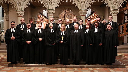 16 junge Geistliche stehen in drei Reihen im Schleswiger Dom. Sie tragen einen Talar und wurden kurz vorher von Bischöfin Nora Steen (blonde Frau in der Mitte) in ihren Dienst als Pastor:innen berufen und gesegnet. 