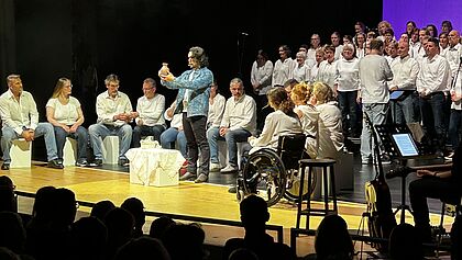 Eine große Bühnenaufführung mit vielen Personen in weißen Oberteilen. Die Menschen vorne stellen das letzte Abendmahl von Jesus in Jerusalem vor. Im Vordergrund steht eine Person und spricht zu einer sitzenden Gruppe, während im Hintergrund ein großer Chor auf der Bühne steht. Seitlich sind Musikinstrumente und technisches Equipment zu sehen; Zuschauer sitzen im dunklen Saal.