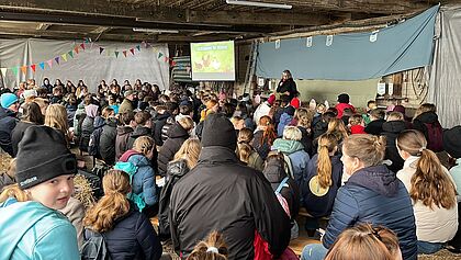 Viele Menschen von hinten fotograniert sitzen in einer Scheune und schauen auf die Sprecherin auf einer Bühne