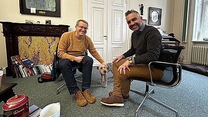 Zwei Männer sitzen sich gegenüber, in der Mitte ein Hund