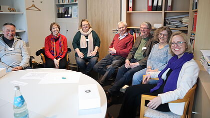 Sieben Personen sitzen an einem Tisch im Gespräch.