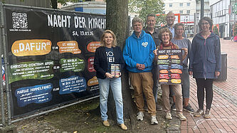 Sieben Personen stehen auf einem gepflasterten Platz neben einem großen Banner zur „Nacht der Kirchen Elmshorn“. Das Banner zeigt Datum, Motto „DAFÜR“, Programmpunkte und einen QR‑Code. Eine Person hält zusätzlich ein Plakat mit denselben Informationen. 