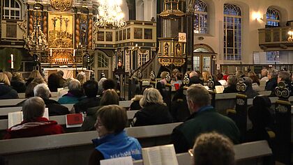 Volle Kirche St. Nikolai. Mitarbeiterin steht am Rednerpult, im Hintergrund der Altar mit der Kreuzigung Jesu. 