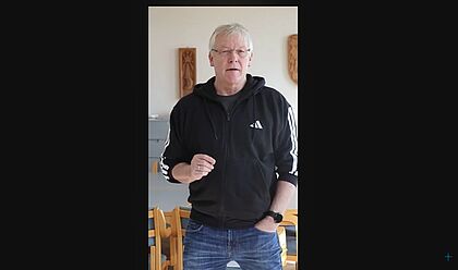 Pastor Jens Siebmann trägt einen schwarzen Hoodie und eine Jeans. Er steht in seinem Esszimmer und sucht eine Nachfolgerin für seine Stelle. Dabei spricht er in die Kamera.