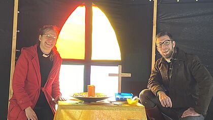 Eine Pastorin und ein Pastor sitzen links und rechts neben einem als Altar gestalteten Tischchen. Im Hintergrund eine Zeltbahn, die wie ein Kirchenfenster anmutet 