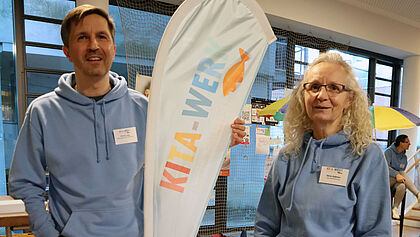Patrick Laas und Dörte Raßmus vom evangelischen Kita-Werk Rantzau-Münsterdorf posieren dem der Kita-Werk Flagge. Sie tragen blaue Hoodies mit Kita-Werk-Logo. 