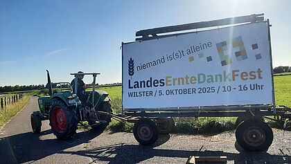 Ein Trekker zieht einen Anhänger. Darauf ist ein Bauzaun an dem wiederum ein Banner gefestigt ist, das für das Landeserntedankfest am 5. Oktober in Wilster wirbt.